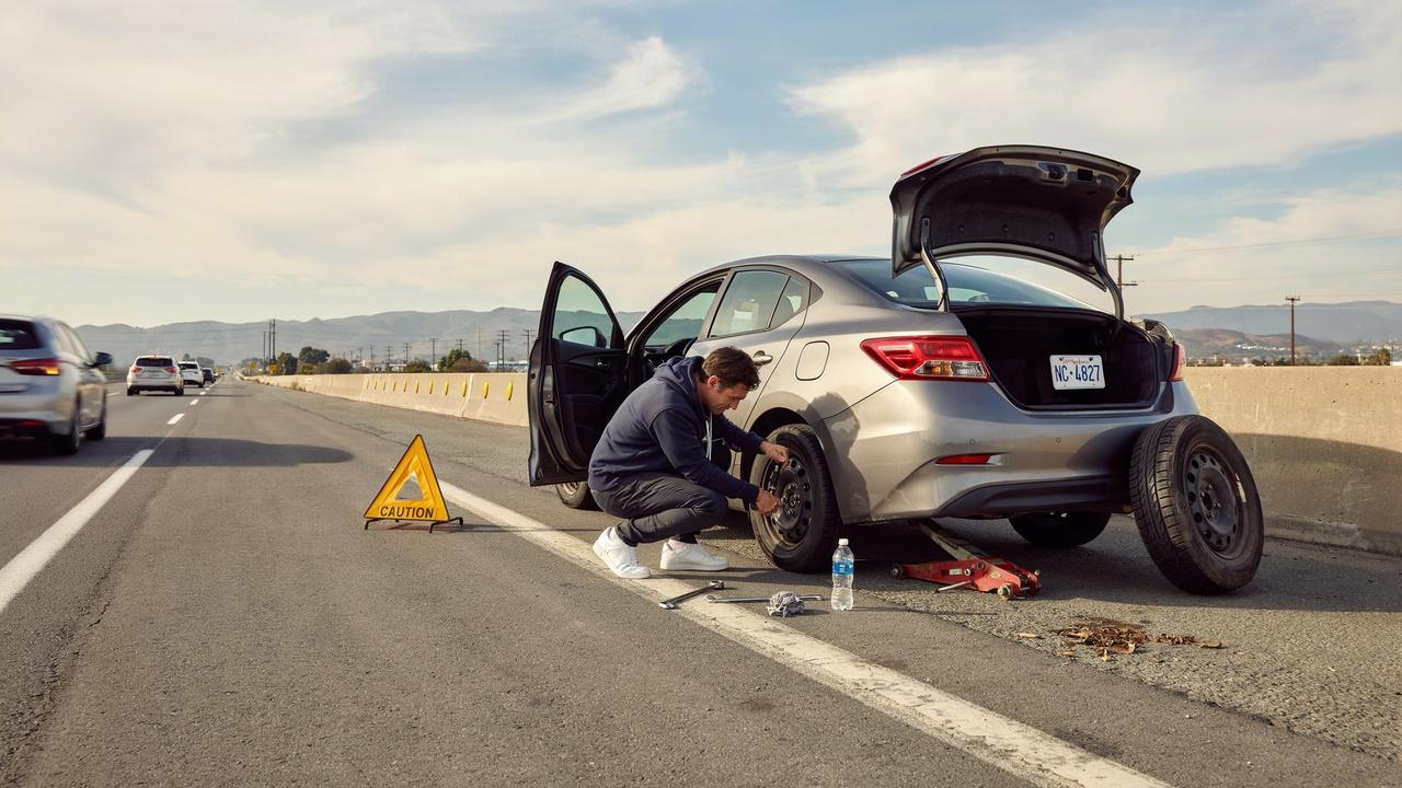 Driver changing a flat tire on the roadside using a spare tire with highway traffic in the background.