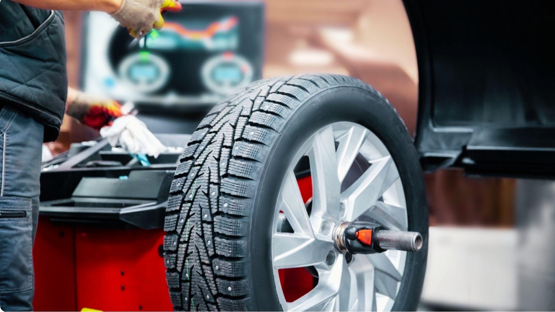 Mechanic balancing a studded winter tire on silver alloy wheel using red tire balancing machine in garage.